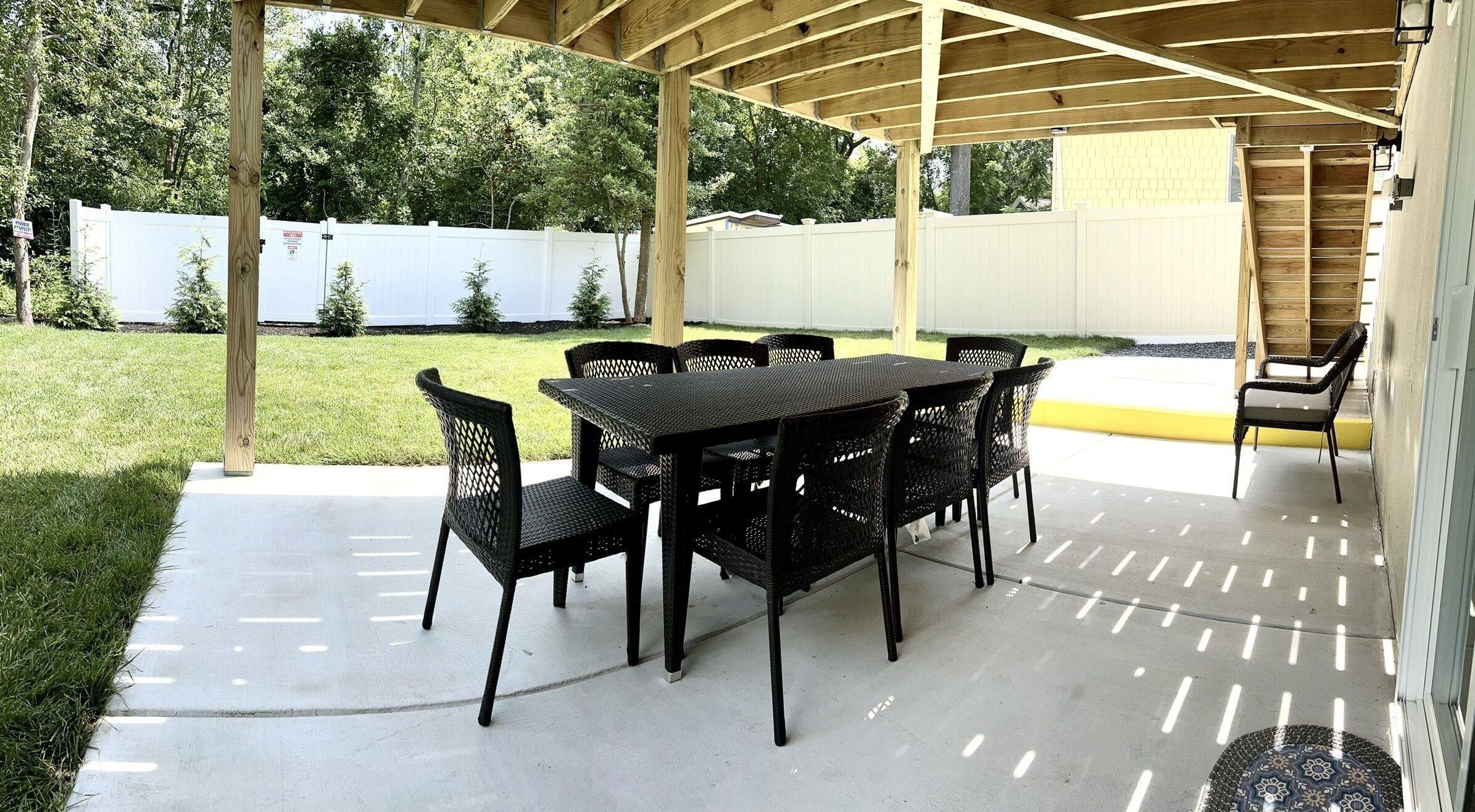 Ein überdachter Patio-Bereich mit Tisch und Stühlen, einem Betonboden und einem weißen Zaun im Hintergrund.