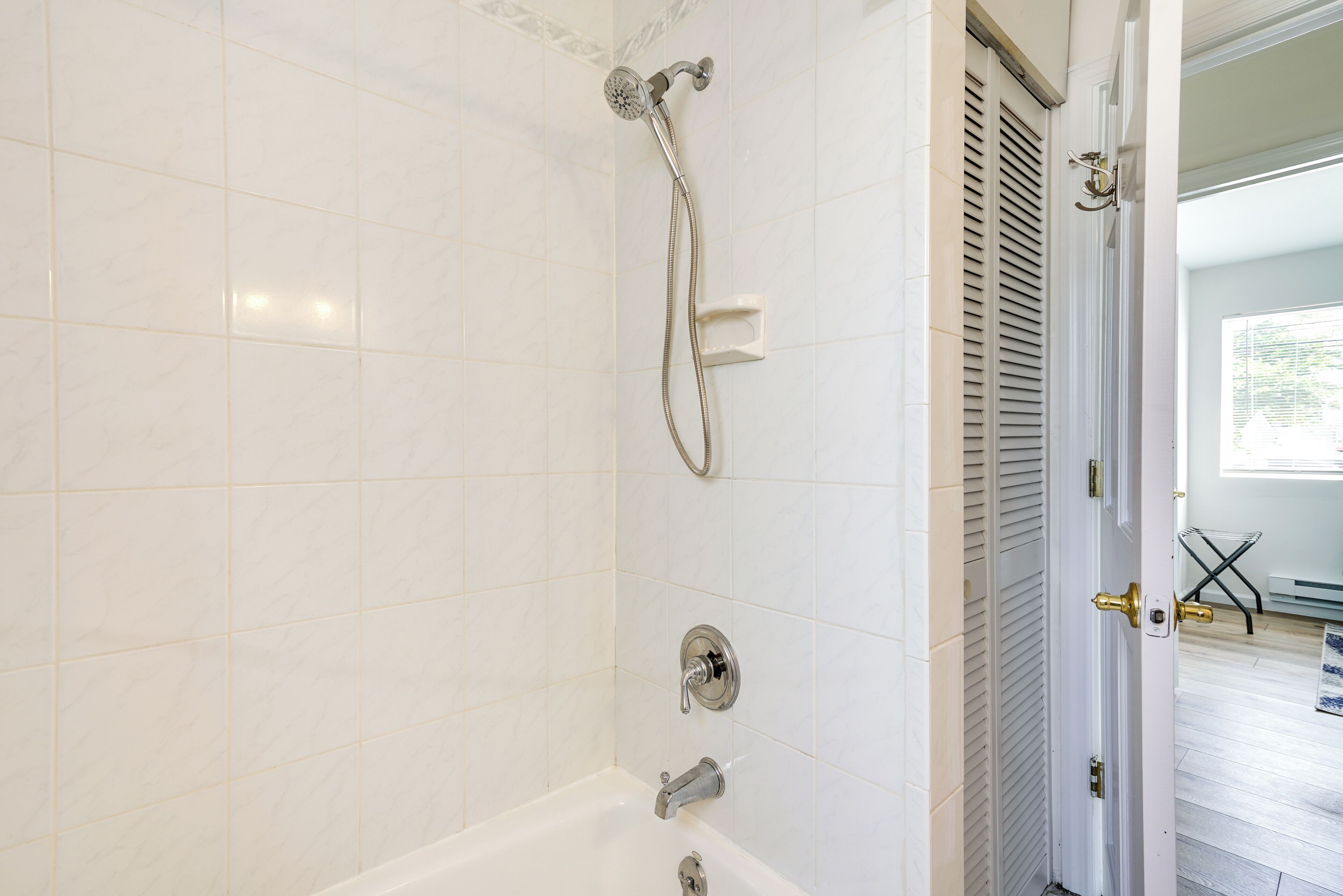 A bathroom with white tiled walls, a bathtub, a showerhead, and a door leading to another room.