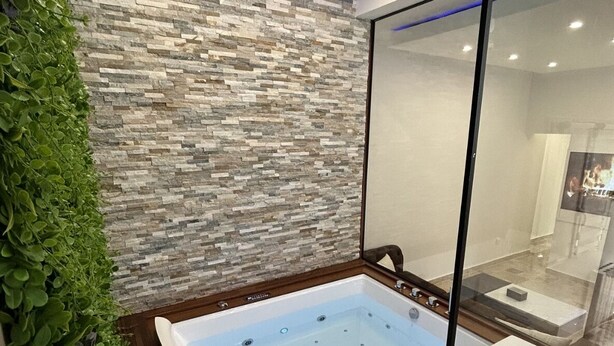 Un baño moderno con pared de piedra, bañera de hidromasaje con mampara de vidrio y pared cubierta de plantas verdes.