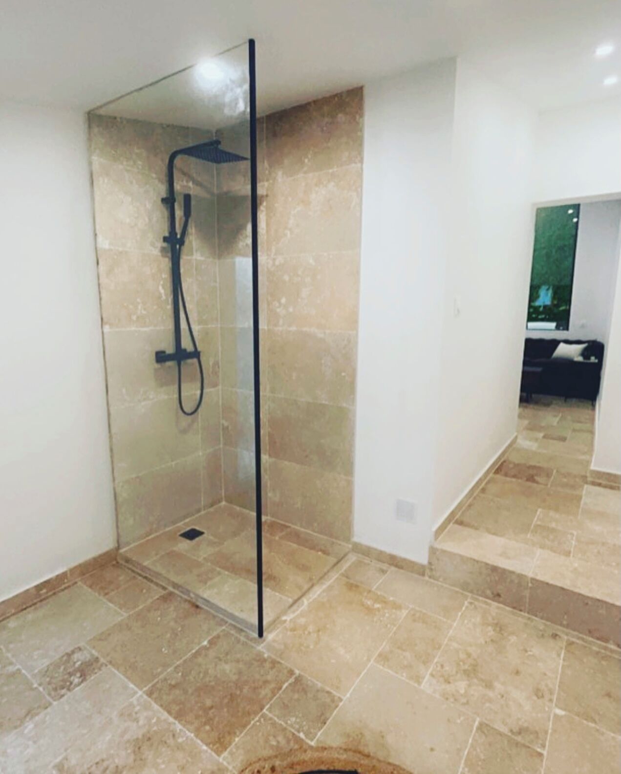 Une douche fermée par des parois de verre, avec un sol carrelé et un drain apparent.