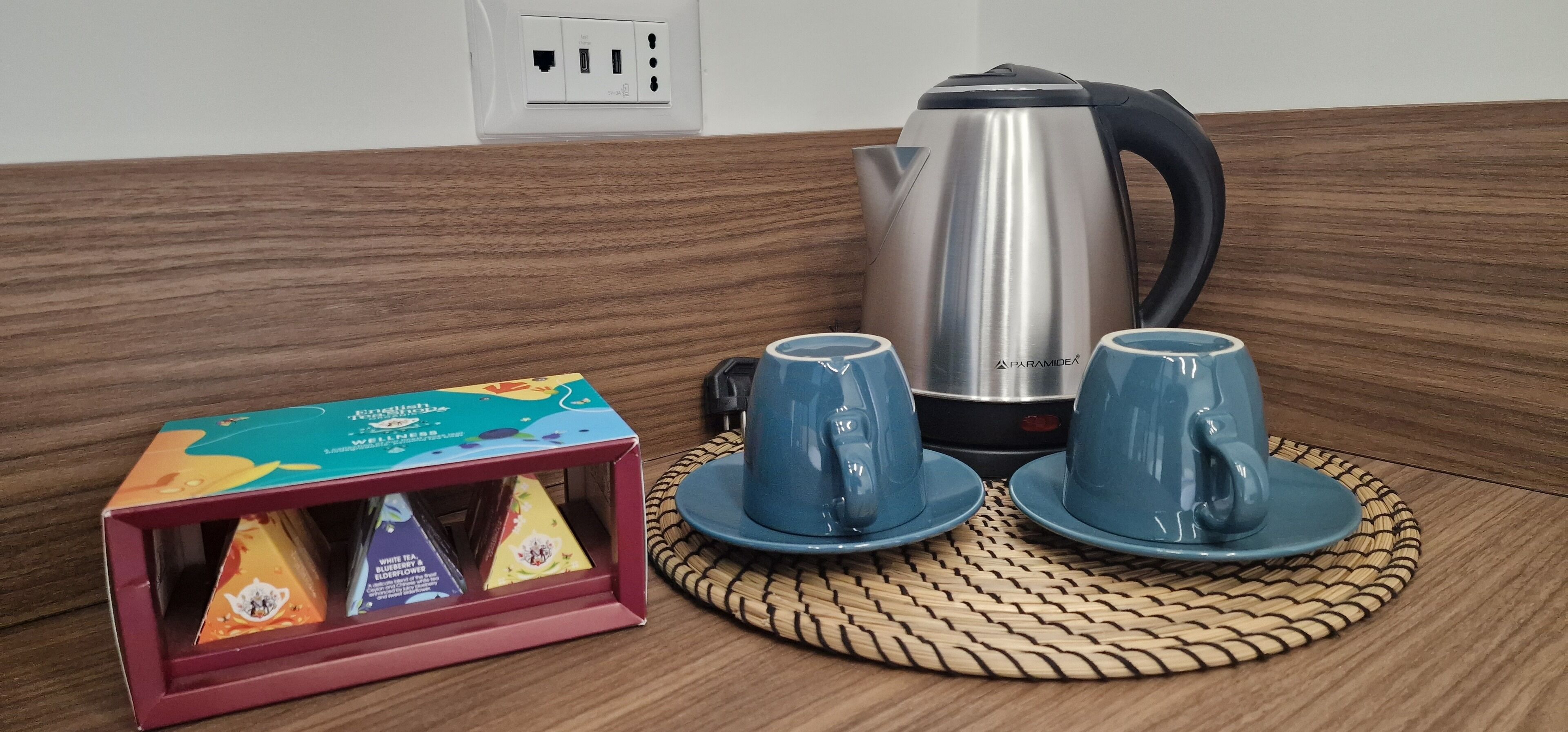 A kitchen area with a stainless steel kettle, two blue cups on saucers, and a box of tea with a colorful design.