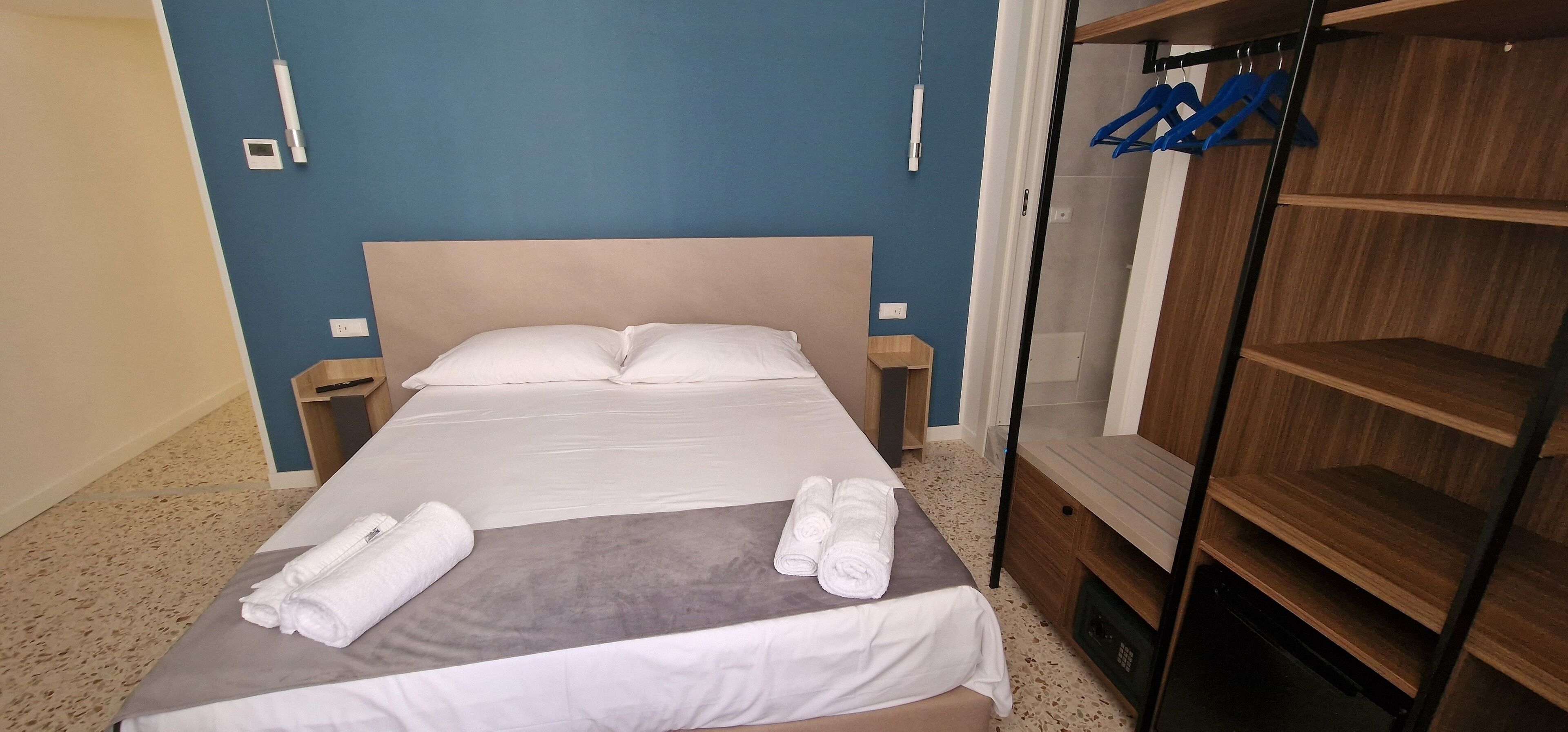 Ein ordentlich bezogenes Bett mit weißen Leinen, einer grauen Decke und aufgerollten Handtüchern seitlich. Zwei Nachttische, einer mit einer Lampe und der andere mit einer Steckdose. Ein Holzschrank mit Spiegel und einem blauen Kleiderbügel befindet sich rechts.