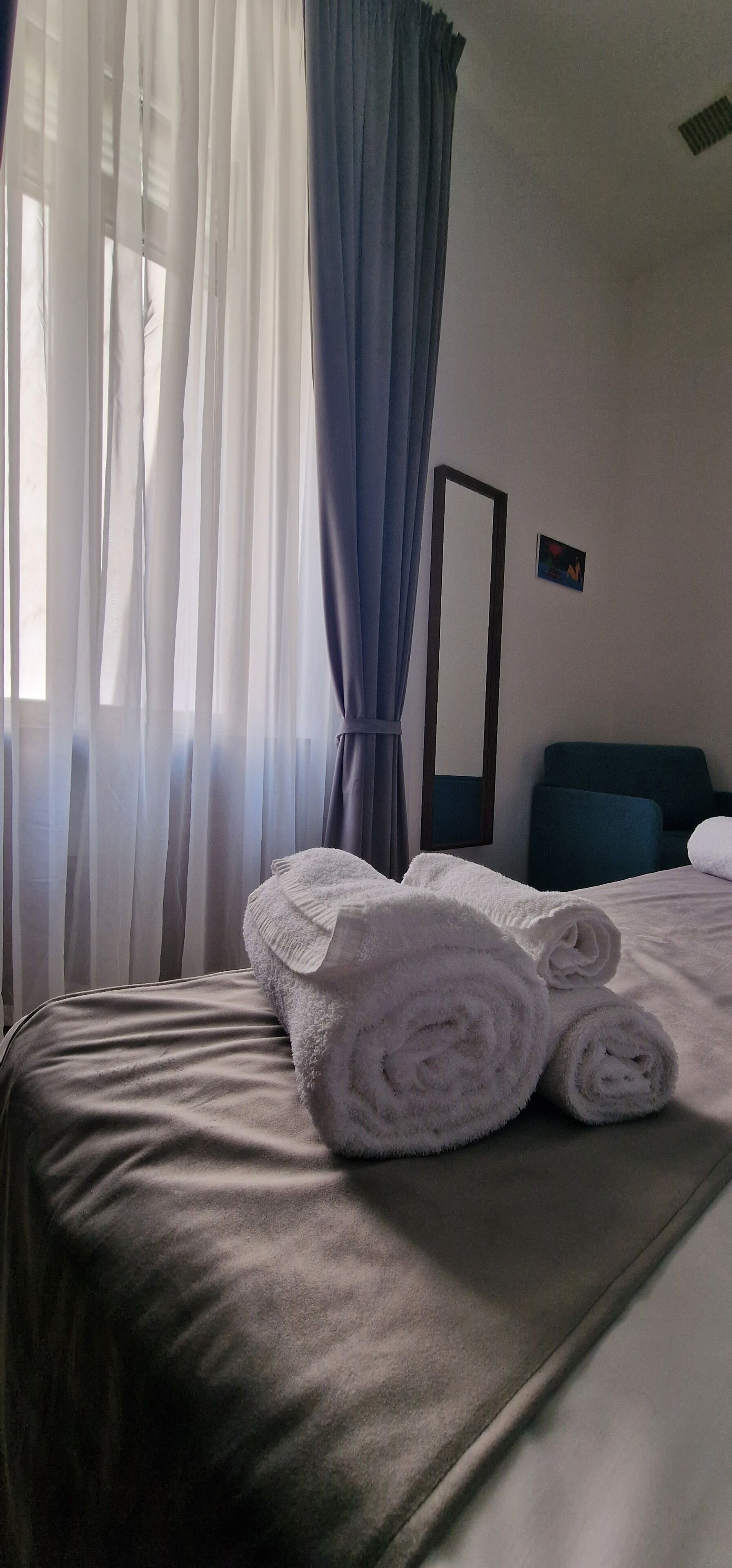 Ein ordentlich bezogenes Bett mit aufgerollten weißen Handtüchern, ein großer Spiegel an der Wand und durchscheinende Vorhänge, die teilweise gezogen sind.