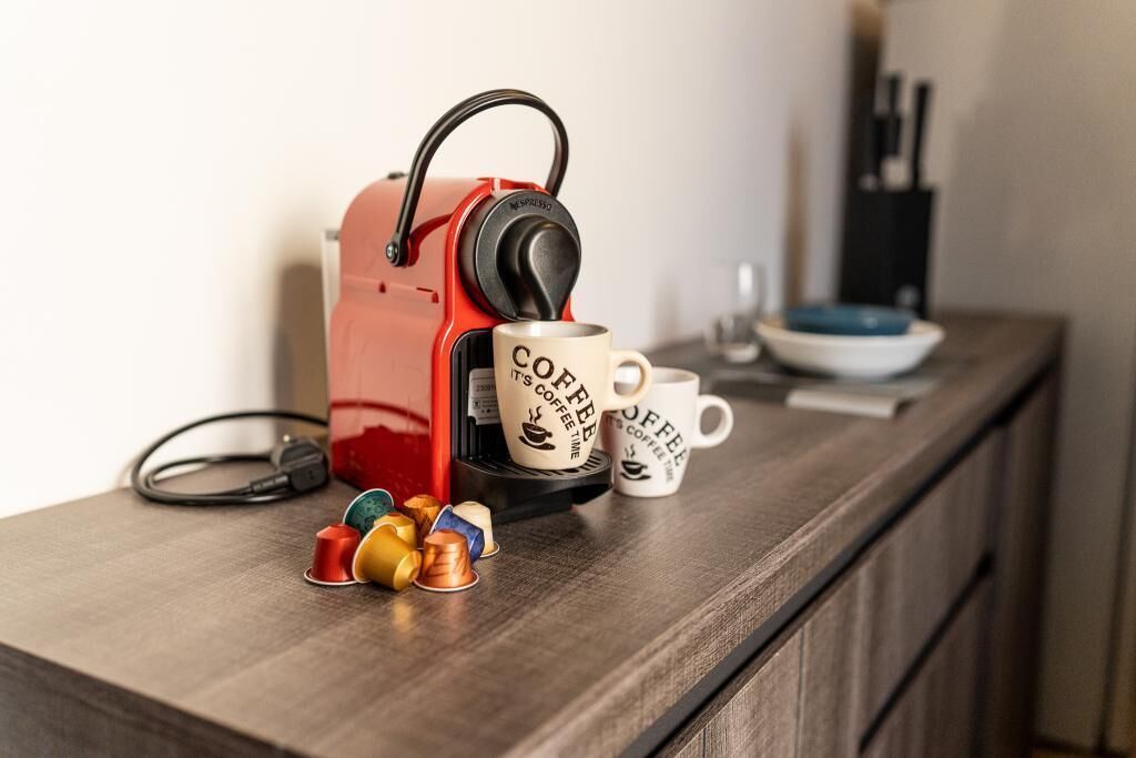 Une machine à café rouge est posée sur un plan de travail en bois, à côté de deux tasses à café et de quelques capsules.