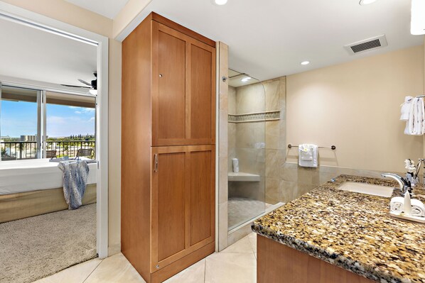 A modern bathroom with a granite countertop, a wooden wardrobe, a glass shower enclosure, and a view of the outdoors.