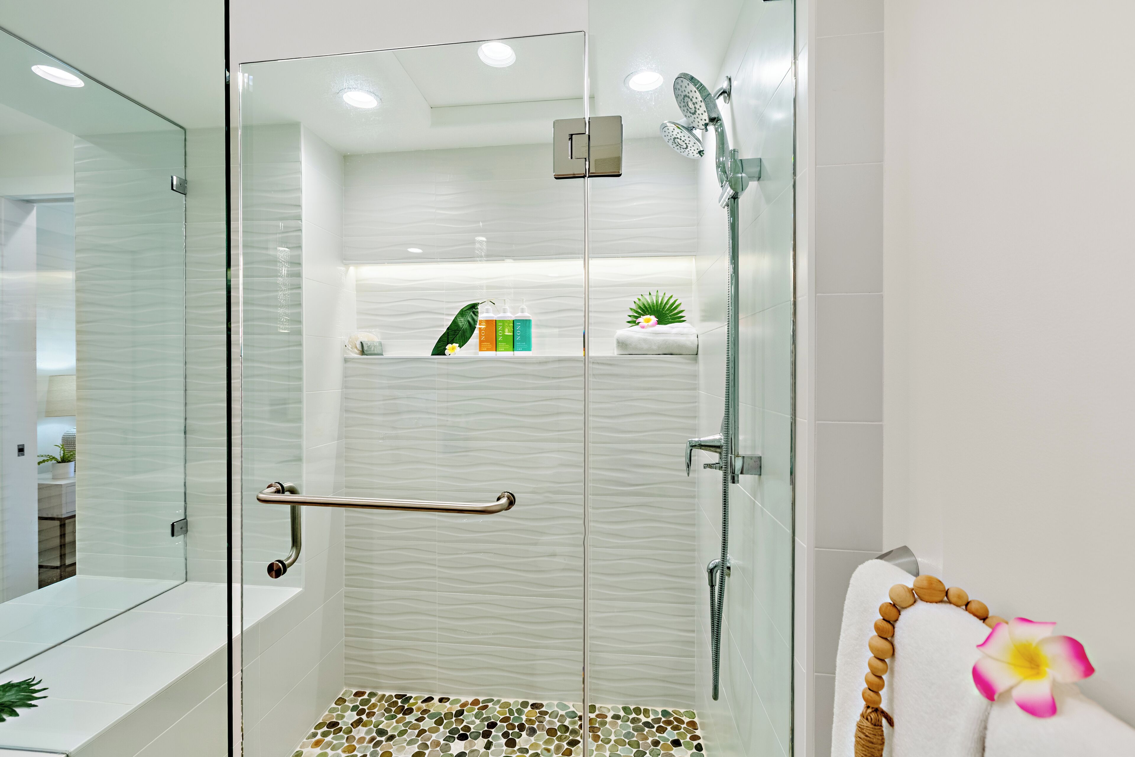 Une salle de bain moderne avec une douche à l’italienne en verre, un mur recouvert de carreaux blancs, une porte en verre, une barre d’appui, un porte-serviettes avec une serviette et une fleur, et une étagère avec des articles de toilette.