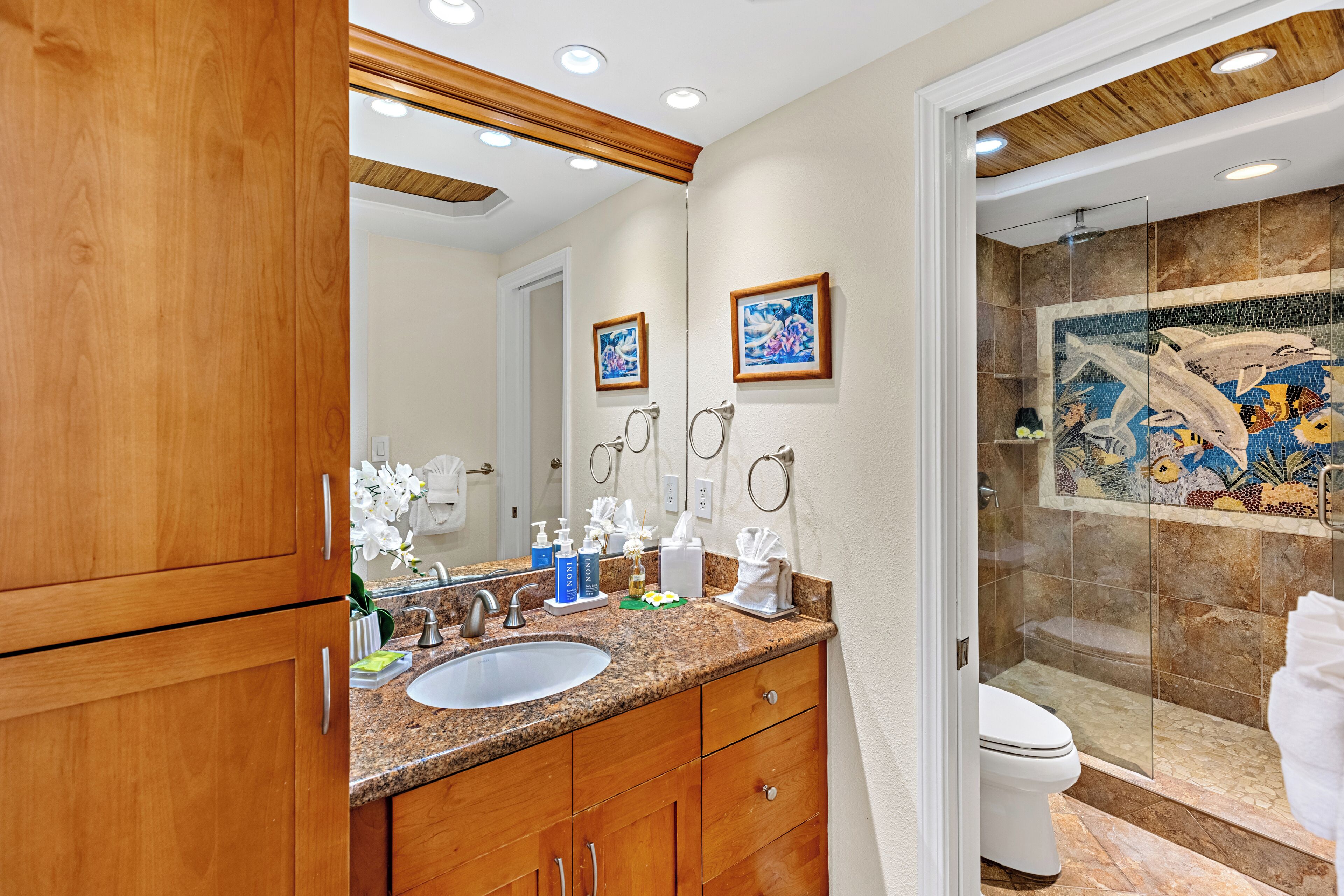 A bathroom with wooden cabinets, a granite countertop, a sink, a toilet, and a shower with mosaic tiles.