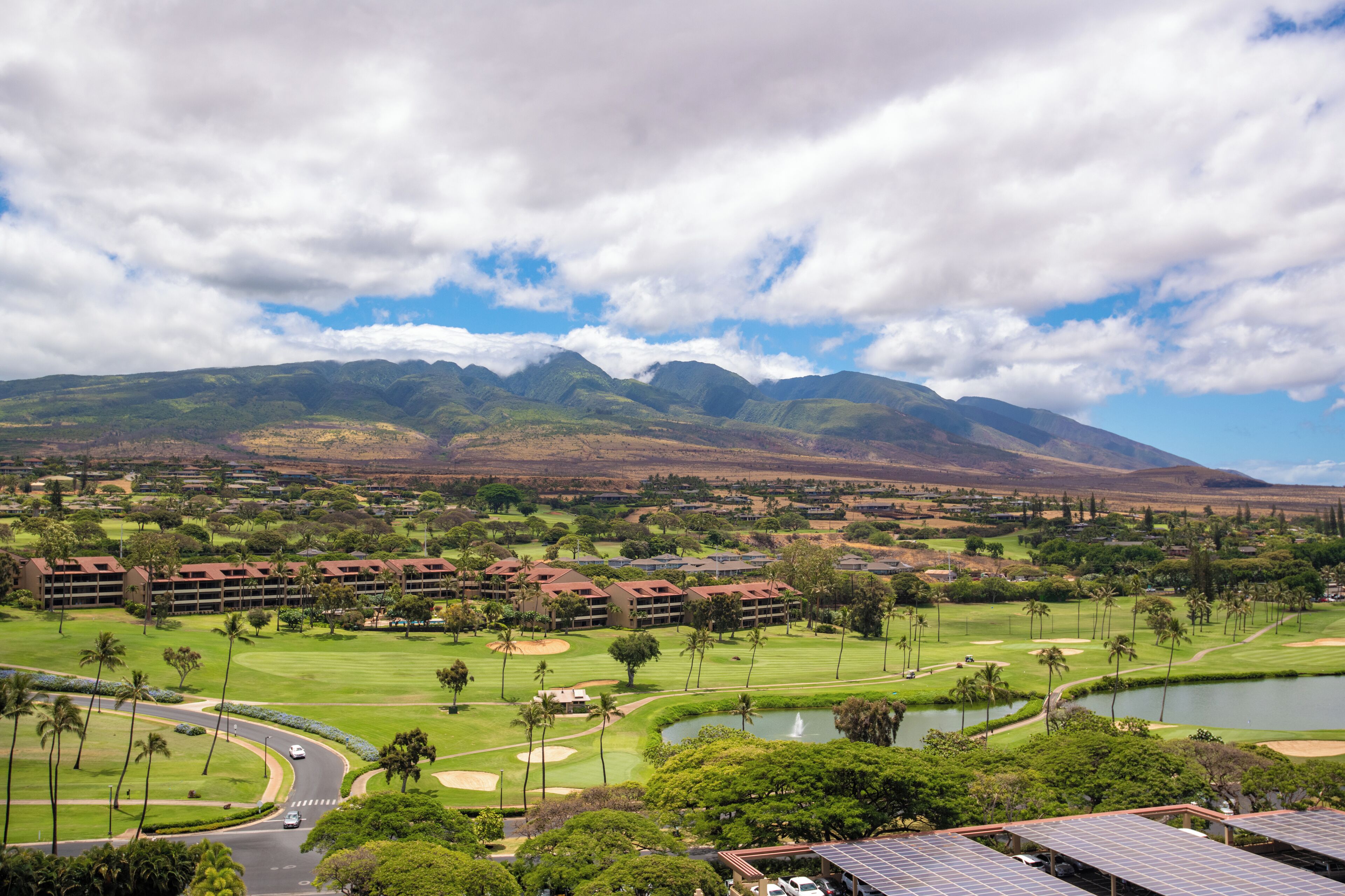 Un campo de golf con vistas a montañas al fondo, una masa de agua y un hotel con varias construcciones.