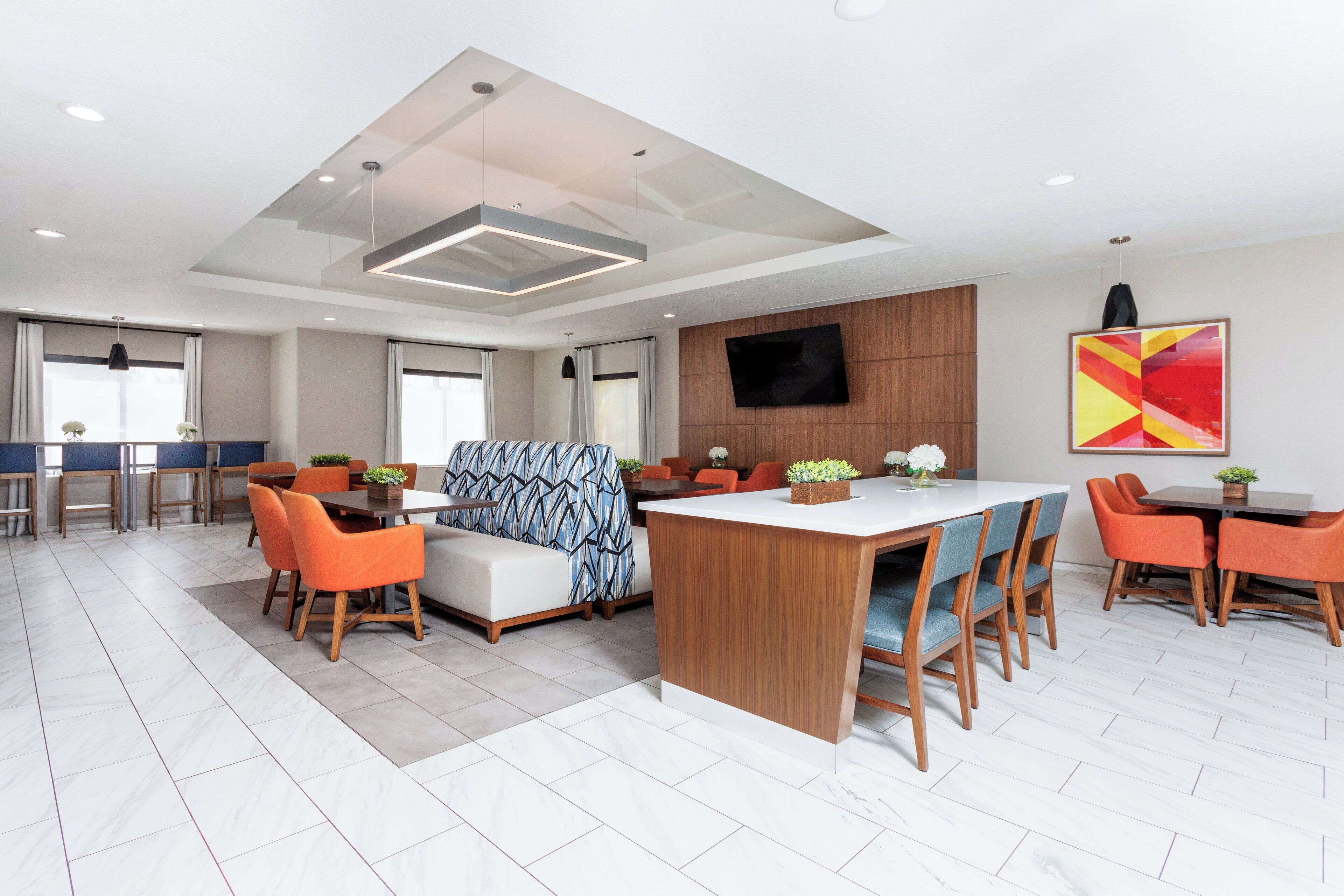 Una habitación de hotel moderna con una isla central, sillas color naranja, un sofá y un televisor montado en la pared.