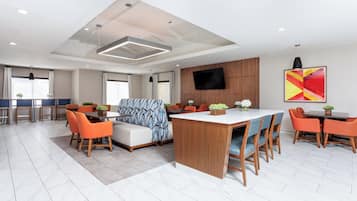 Una habitación de hotel moderna con una isla central, sillas color naranja, un sofá y un televisor montado en la pared.