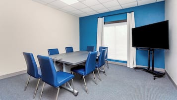 A conference room with a large table, blue chairs, a flat-screen TV, and a window with white curtains.