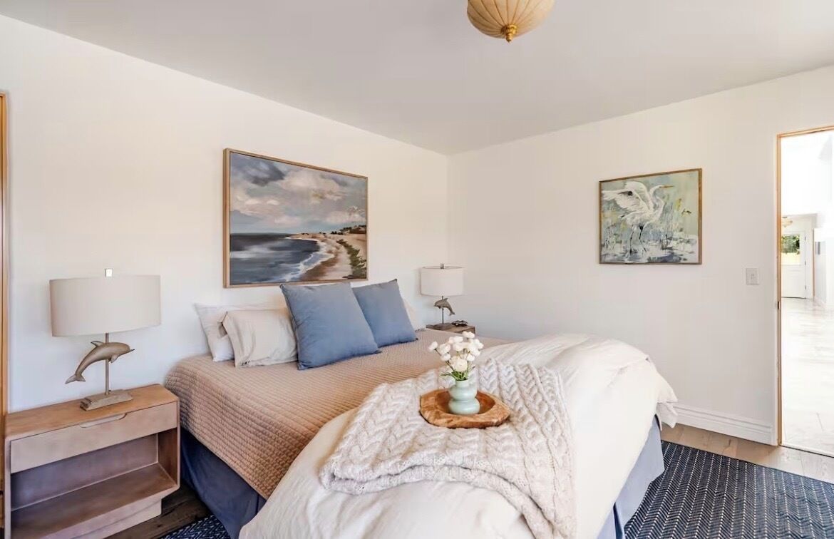 Ein Schlafzimmer mit einem Bett, einem Nachttisch, einer Lampe, einem Strandbild und einer Tür zu einem anderen Raum.