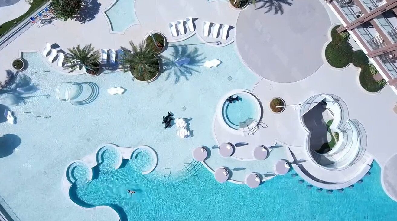 Vista aérea de un área de piscina en un resort, con varias piscinas, sillas de descanso y palmeras.