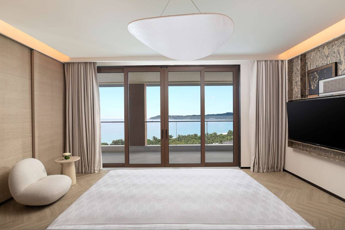 A modern hotel room with a large bed, a seating area, a small table, and a view of the sea.