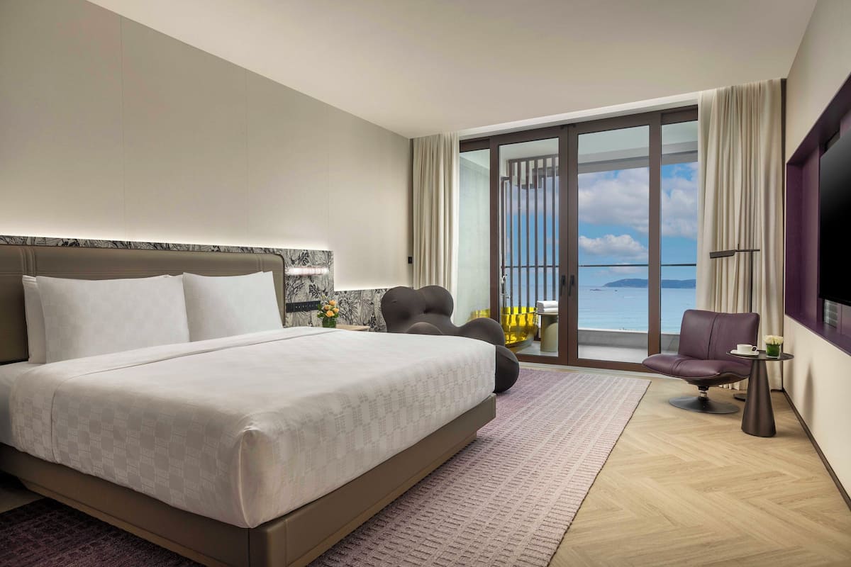 A modern hotel room with a large bed, a seating area with a chair and a small table, and a balcony with a view of the sea.