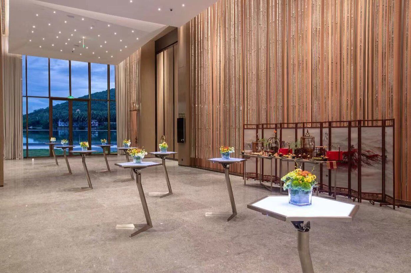 A spacious hotel lobby with multiple tables, a large window offering a view of the outdoors, and a decorative wall with vertical panels.