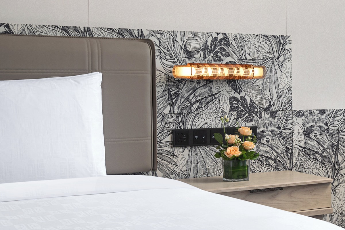 A neatly made bed with a white pillow and a headboard against a wall with a leaf-patterned wallpaper. A bedside table with a vase of flowers and a lamp is visible.