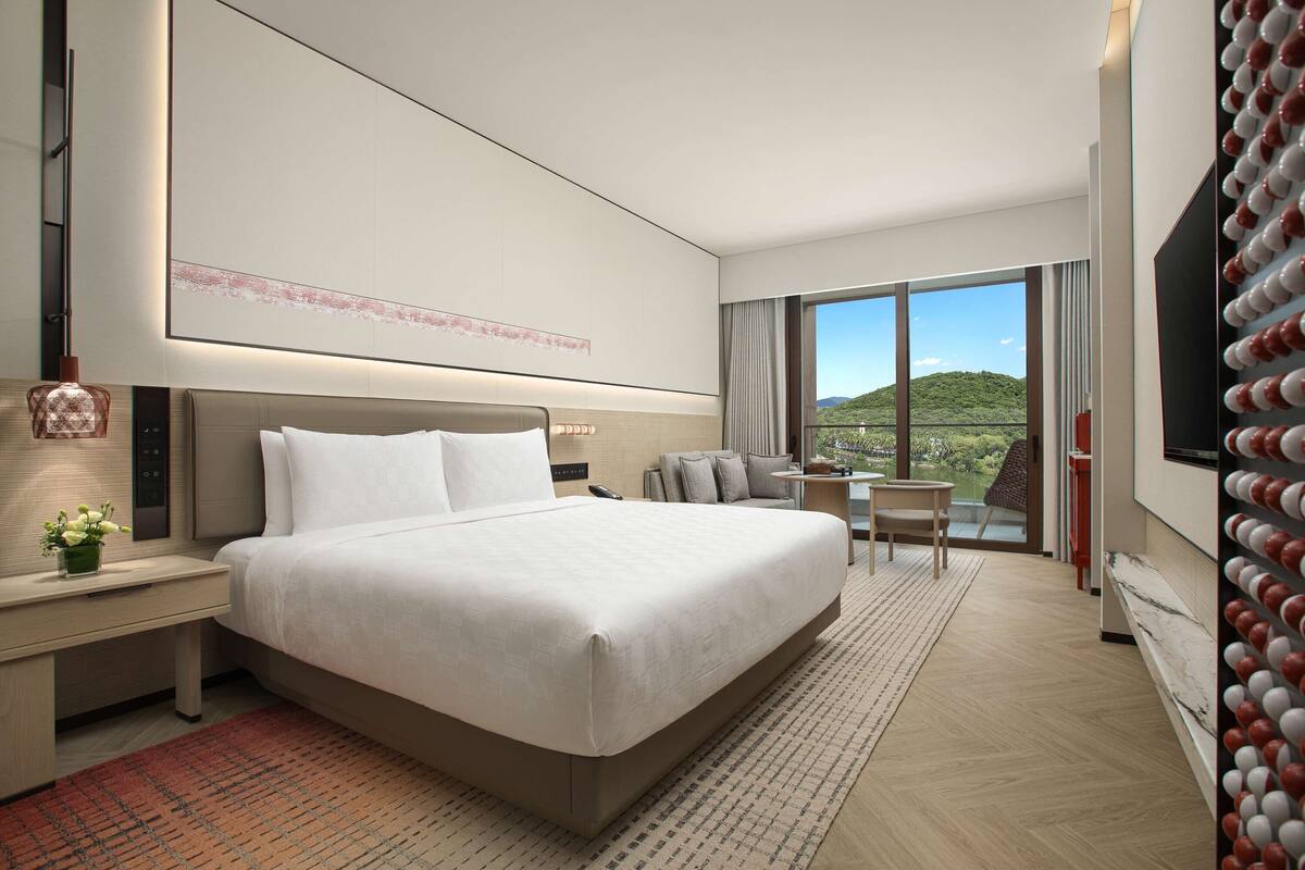 A modern hotel room with a large bed, a desk with a lamp, a small table with chairs, and a large window offering a view of the outdoors.