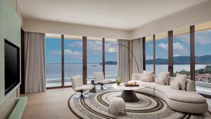 Un salon moderne avec un grand canapé en forme de L, une table basse et un téléviseur fixé au mur. La pièce est dotée de grandes fenêtres offrant une vue sur la mer et les montagnes.