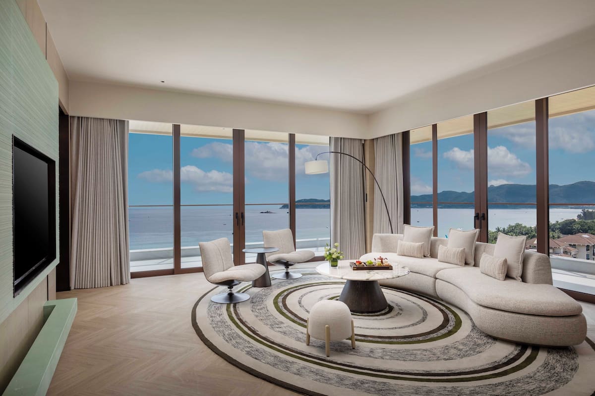 A modern living room with a large L-shaped sofa, a coffee table, and a TV on the wall. The room has large windows with a view of the sea and mountains.