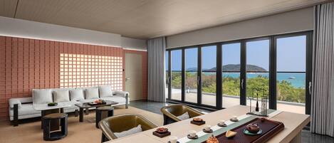A modern hotel room with a large dining table set for tea, a sofa, and a view of the sea.