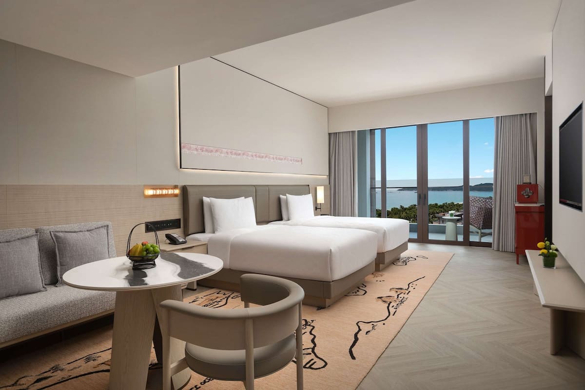 A hotel room with a large bed, a sofa, a small table with a fruit basket, and a view of the sea through large windows.