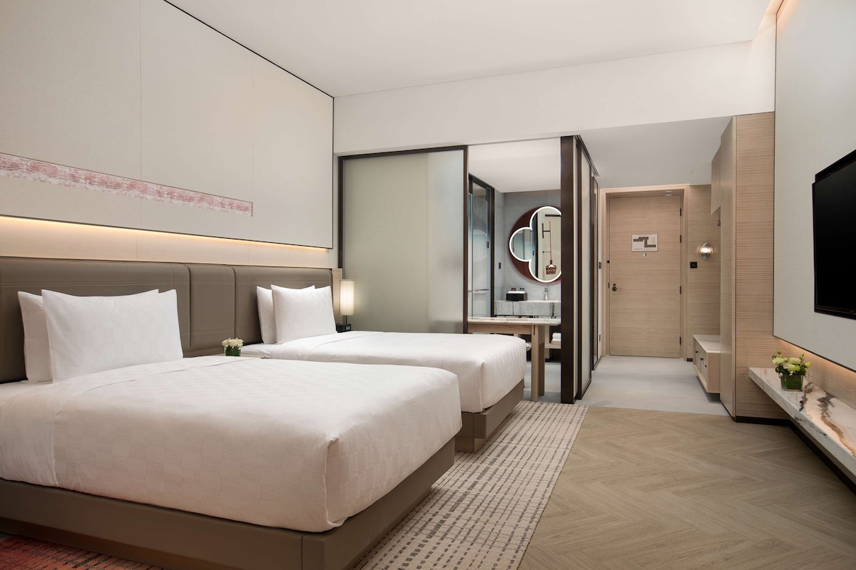 A hotel room with two beds, a large mirror, a TV, and a small table with a vase of flowers.
