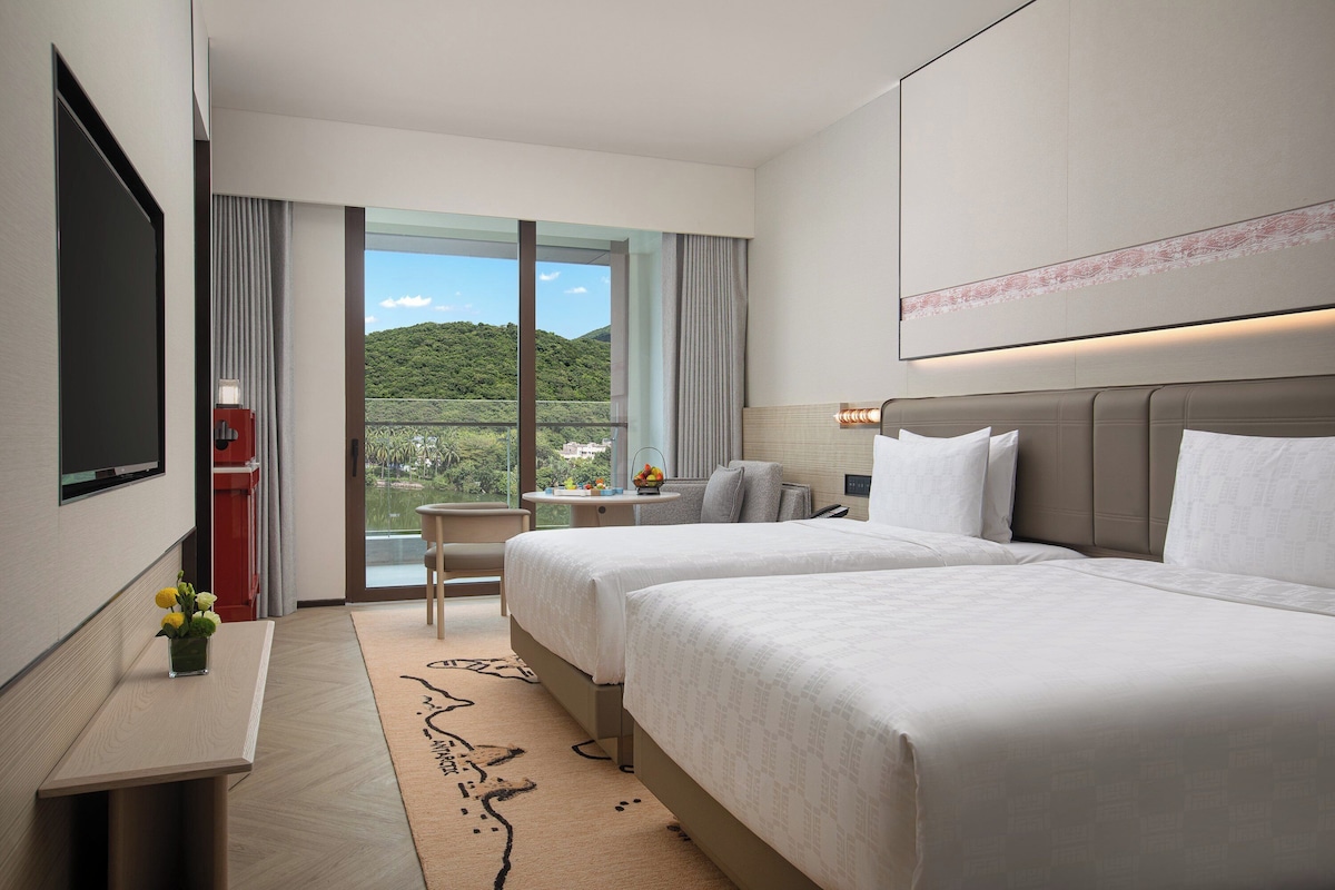 A hotel room with two beds, a desk, a TV, and a view of a green landscape.