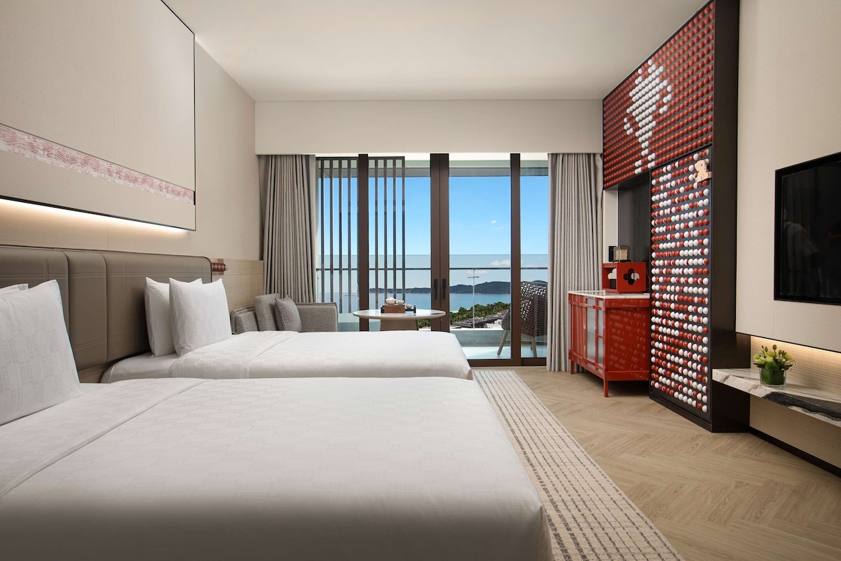 A modern hotel room with two beds, a large window with a view, a red decorative wall, and a flat-screen TV.