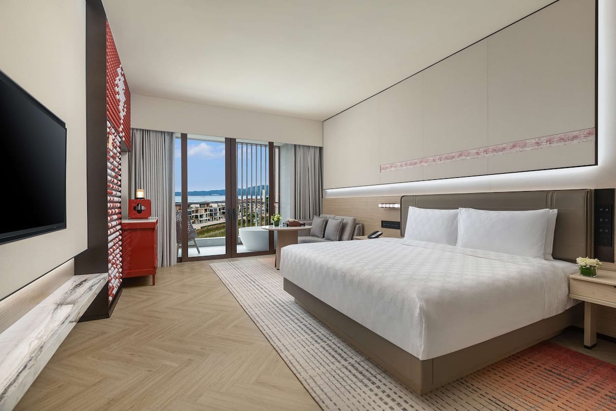 A modern hotel room with a large bed, a flat-screen TV, a red accent wall, a large window with a city view, and a balcony with a view of the city.