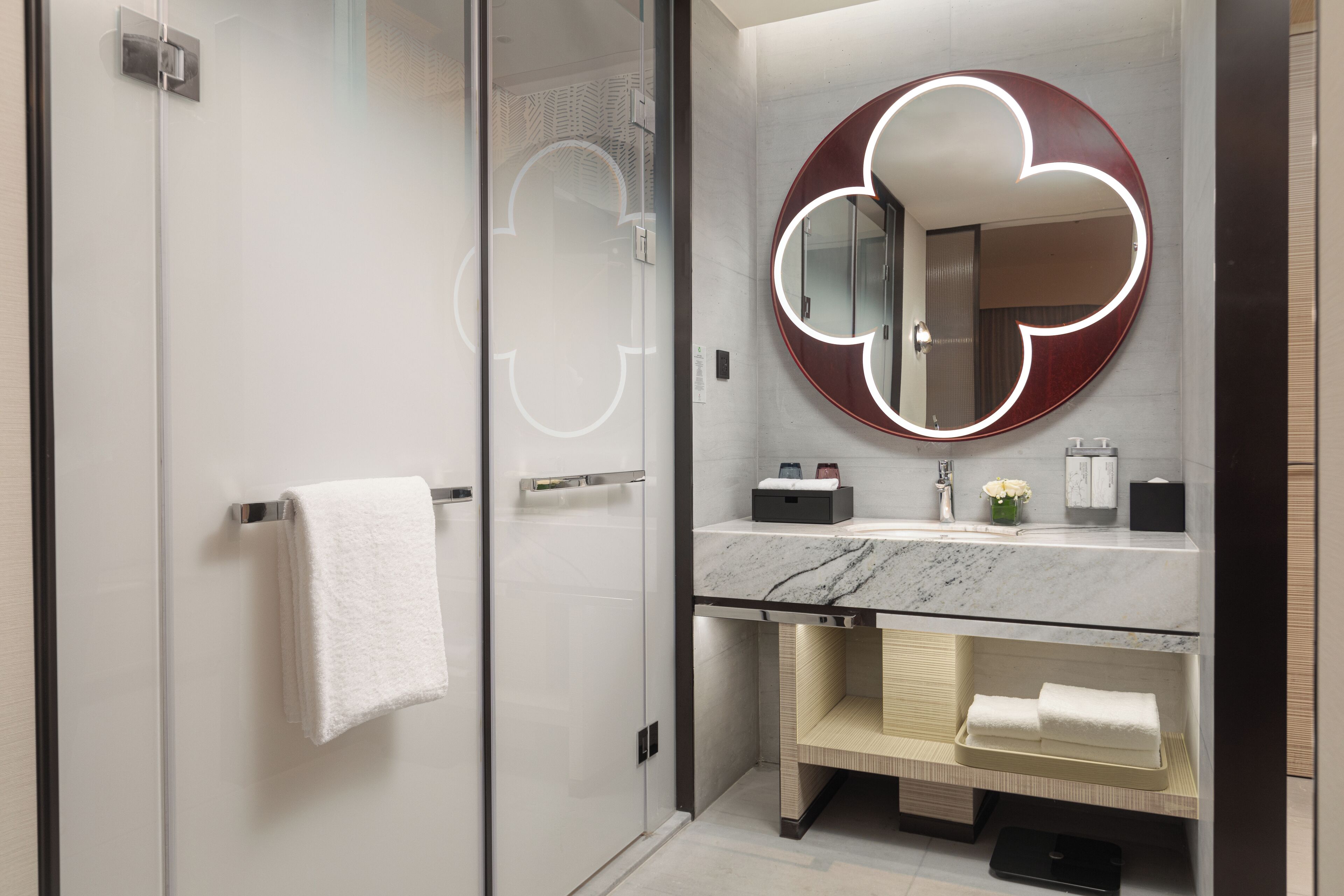 A modern bathroom with a glass shower enclosure, a marble sink, and a round mirror with a light halo.
