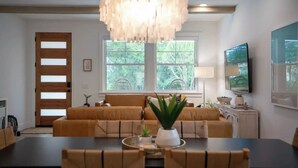 A modern living room with a large sofa set, a dining table with chairs, a chandelier, and a television mounted on the wall.