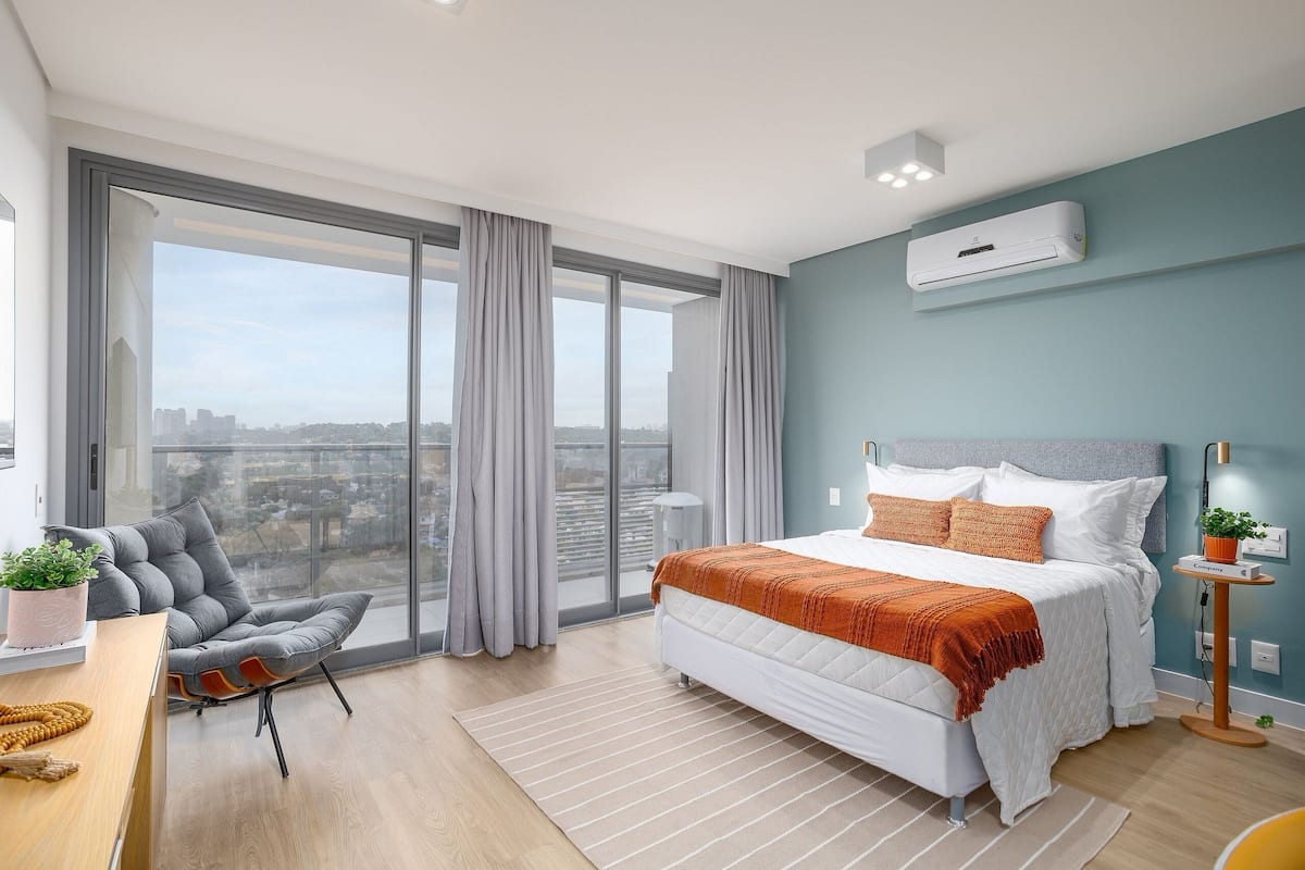 A modern bedroom with a large bed, a grey upholstered chair, a desk with a plant, and a city view through large windows.