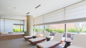 A modern lounge area with sun loungers, a wooden bench, and a large window offering a view of greenery. - Parque Jockey Pinheiros (São Paulo)