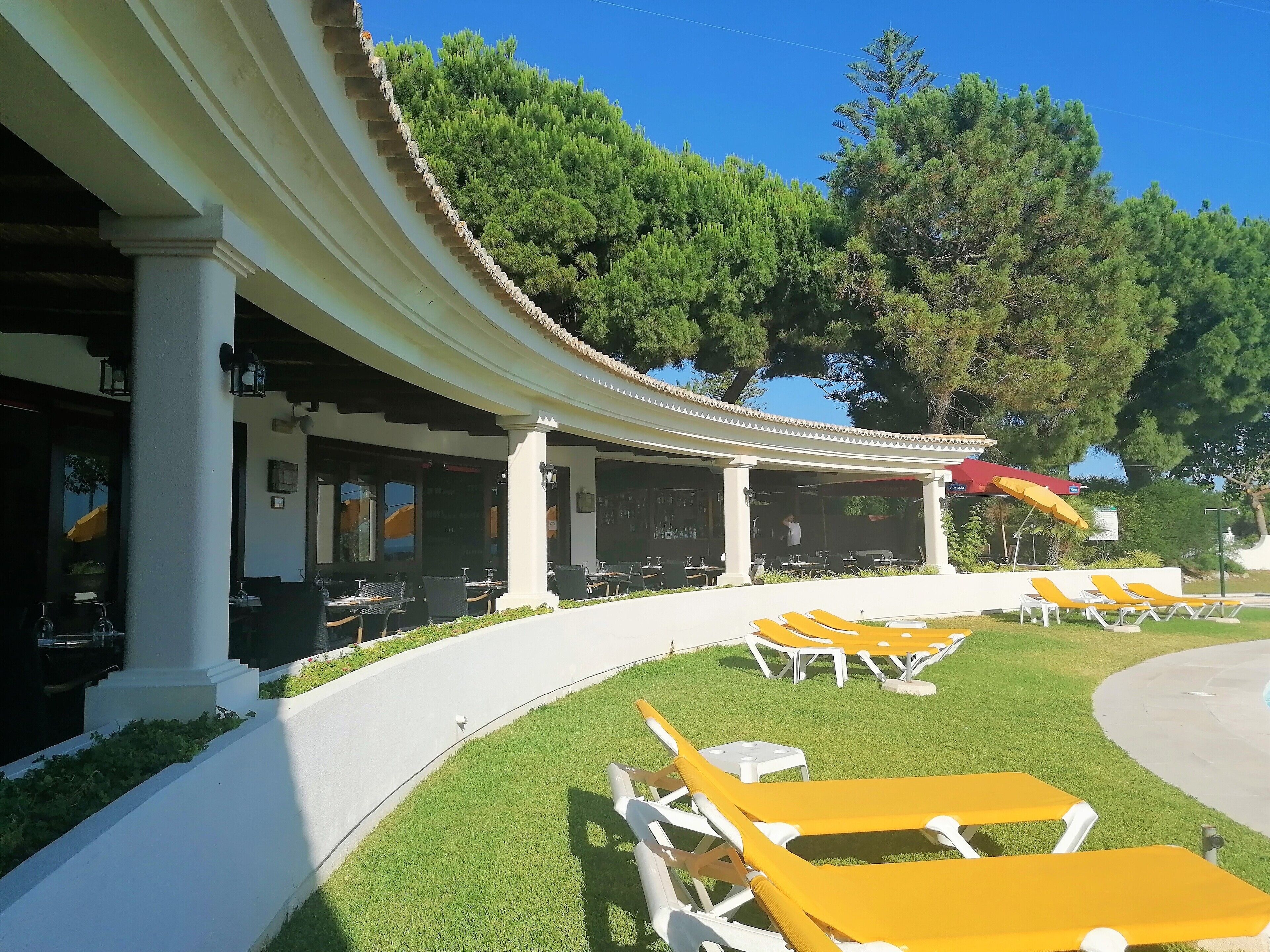 Un'area relax all'aperto coperta, con tavoli e sedie, un solarium con poltrone gialle e un edificio con colonne e una pergola.