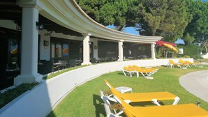 Un área de estar al aire libre cubierta con mesas y sillas, un espacio de descanso junto a la piscina con sillas amarillas, y un edificio con columnas y una pérgola.
