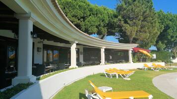 Un'area relax all'aperto coperta, con tavoli e sedie, un solarium con poltrone gialle e un edificio con colonne e una pergola.