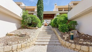 Una escalera de piedra conduce a la entrada de un edificio, bordeada de vegetación y un sendero adoquinado.