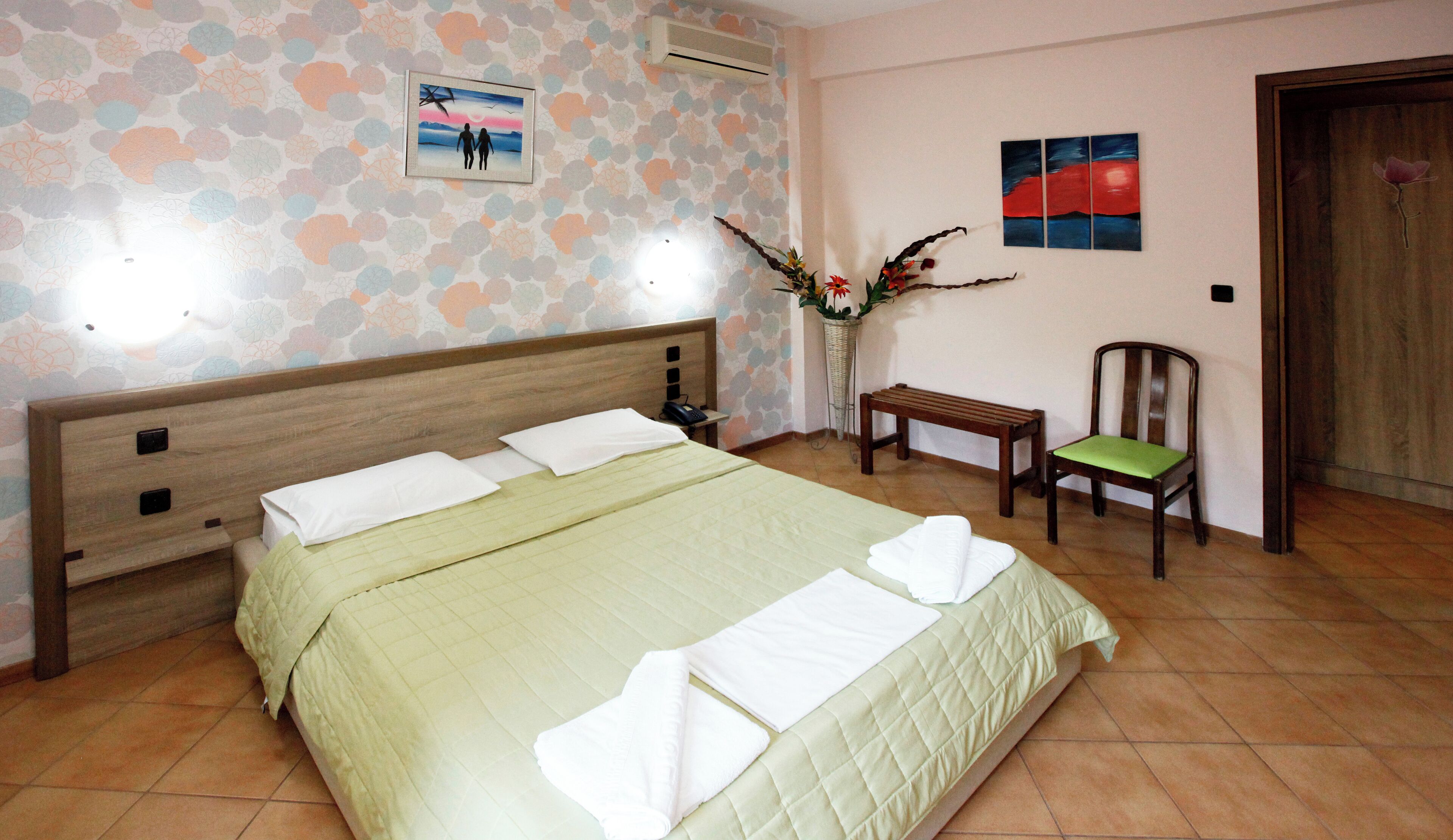 A neatly made bed with a green bedspread and white pillows, a wooden headboard, a bedside table with a phone, a chair with a green cushion, a wall with a floral wallpaper, and a painting of a sunset.