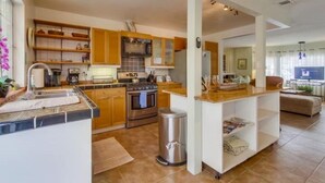 Una cocina moderna con armarios de madera, un lavamanos de acero inoxidable y una isla central. Hay un cubo de basura y una pequeña zona de estar con una mesa y sillas.