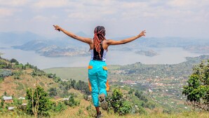 A person standing on a hill with arms outstretched, overlooking a valley with houses and a body of water. - Frama Nature Adventure (Rugarama)