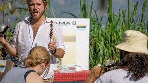 A person holding a stick with food, standing in front of a table with bottles and a sign. - Frama Nature Adventure (Rugarama)