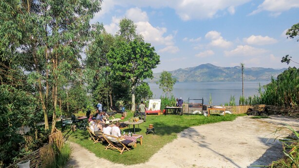 A serene outdoor seating area with tables and chairs, surrounded by lush greenery and overlooking a calm lake with distant mountains. - Frama Nature Adventure (Rugarama)