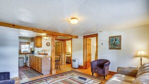 A spacious living room with a large sofa, a coffee table, and a fireplace. There is a kitchen area with wooden cabinets and a dining table. The room has a high ceiling with exposed beams and a large window providing natural light. - Secluded Stonewall Cabin w/ Mountain View! (Weston)