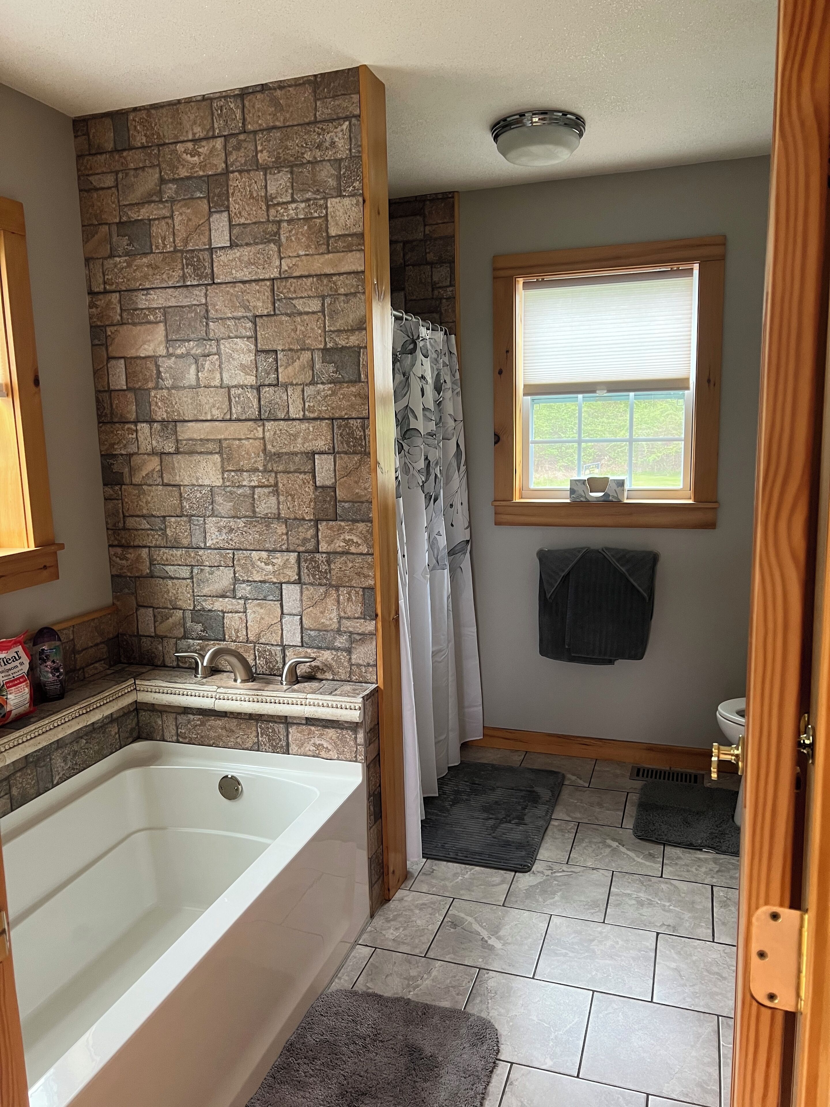 A bathroom with a stone wall, a bathtub, a window with blinds, and a shower curtain.