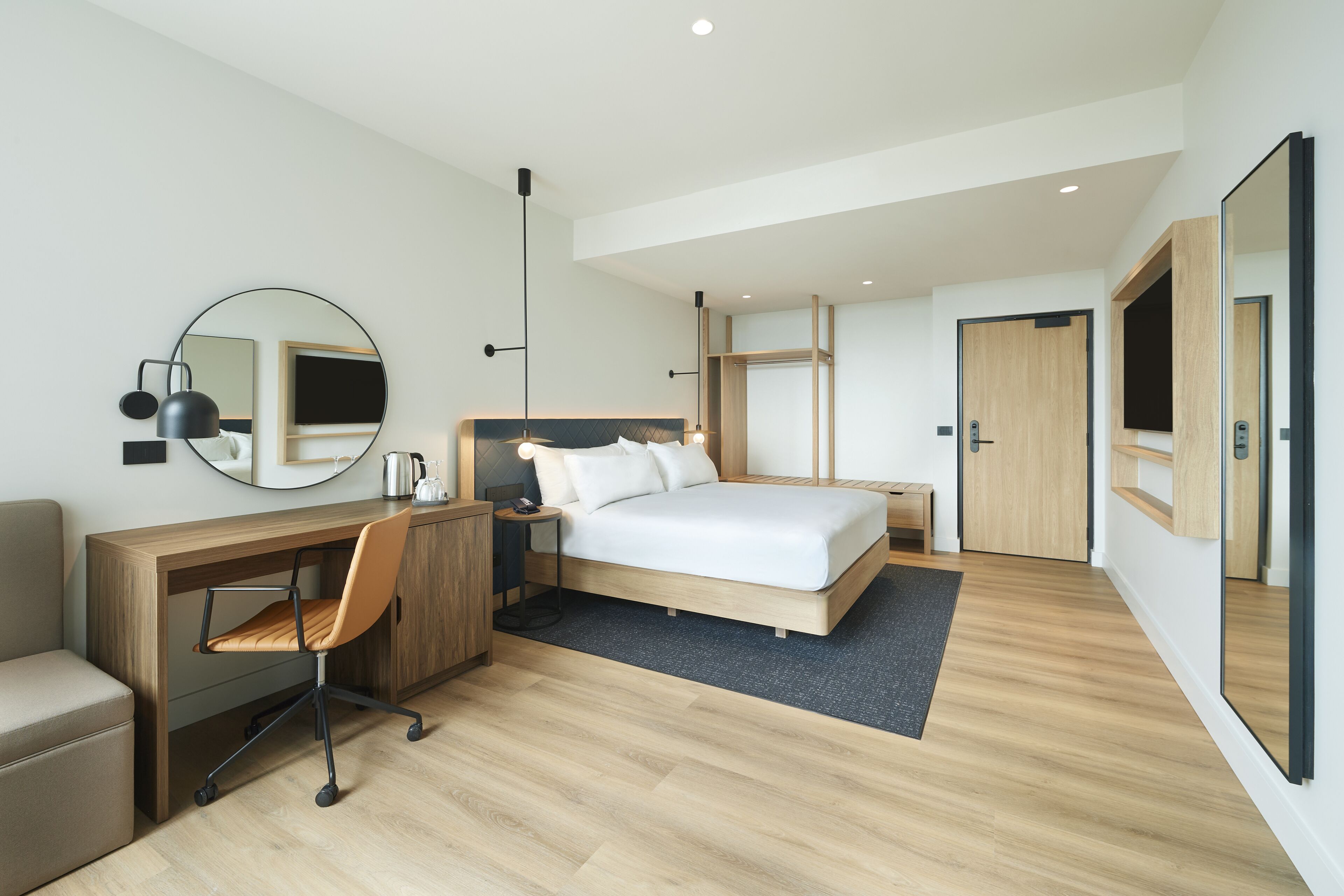 Ett modernt hotellrum med ett skrivbord i trä, en bekväm stol, en säng med vita sängkläder, en rund spegel och ett stort fönster.