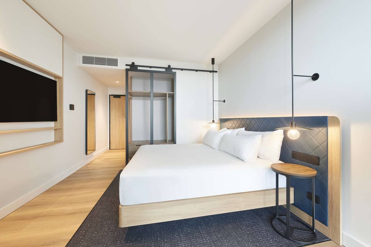 Una habitación de hotel moderna con una cama grande, una mesita de noche, un televisor montado en la pared y un ventilador de techo.