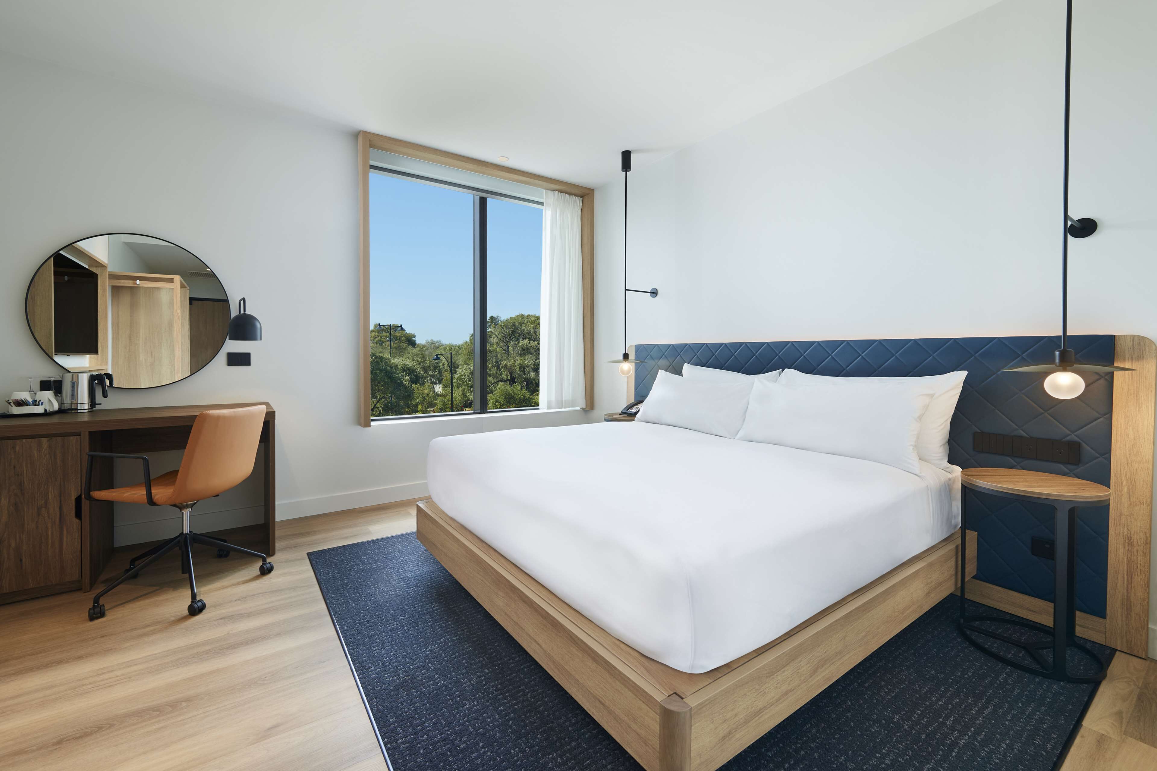 A modern hotel room with a large bed, a desk with a chair, a round mirror, and a view of the outdoors through a large window.