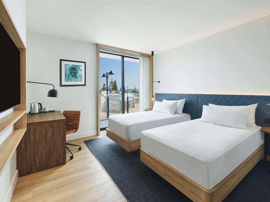 A modern hotel room with two beds, a desk, and a large window offering a view of the city.