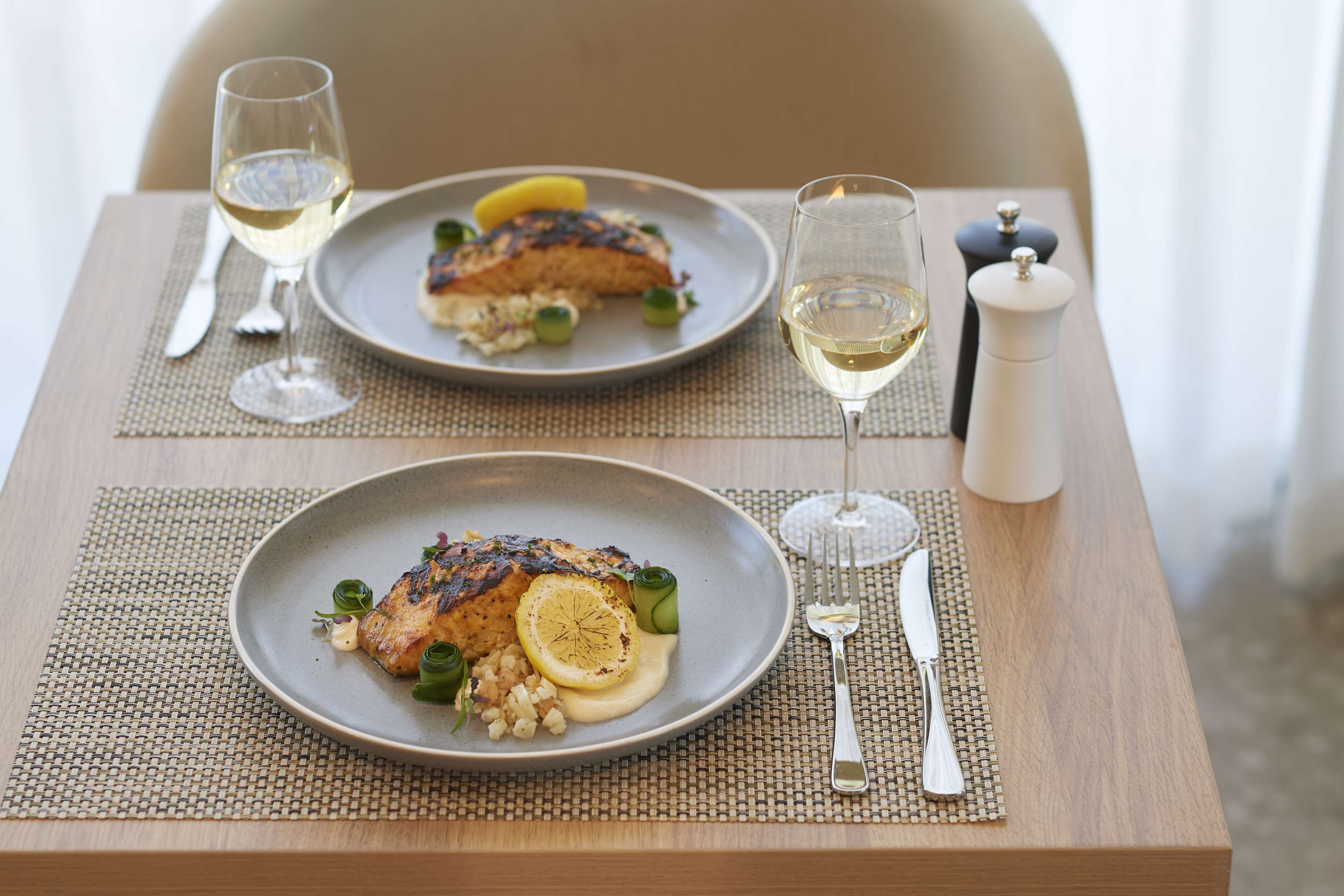 Una mesa de comedor con dos platos de comida, cada uno con una copa de vino blanco y un salero y pimentero.